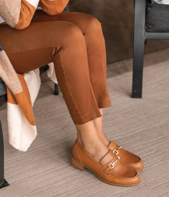 MUNRO TAN LEATHER GRYFFIN LOAFER WITH SADDLE & METAL ORNAMENT  ON MODEL WEARING TAN SLACKS AND COLORFUL SWEATER SITTING IN GRAY CHAIR WITH COFFEE CUP IN HAND-LIFESTYLE